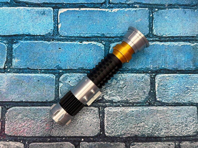 May include: A replica lightsaber with a silver, gold, and black design. The hilt has a black grip with circular indentations. The top and bottom are silver, with a gold section in the middle. The lightsaber is a prop.
