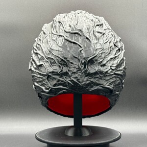 Puede incluir: Una esfera negra impresa en 3D con una superficie texturizada que se asemeja a un cerebro. La esfera est&aacute; sobre un soporte negro con una base roja.