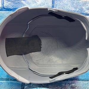 May include: A gray helmet interior with a black fabric strap and a metal frame. The helmet has a smooth, curved design and is likely made of plastic. The interior shows circular patterns.