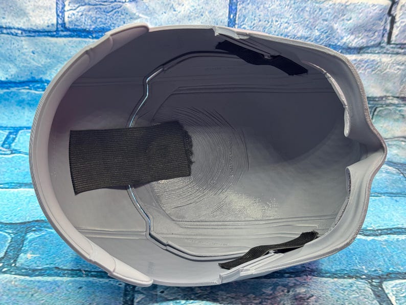 May include: A gray helmet interior with a black fabric strap and a metal frame. The helmet has a smooth, curved design and is likely made of plastic. The interior shows circular patterns.