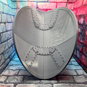 May include: A gray, heart-shaped shield with riveted metal plates. The shield has a curved design and is set against a brick wall background. The shield is a prop or decorative item.