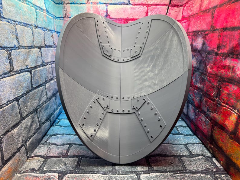 May include: A gray, heart-shaped shield with riveted metal plates. The shield has a curved design and is set against a brick wall background. The shield is a prop or decorative item.