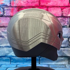 May include: A silver and black helmet with a brushed metal finish, displayed on a black stand. The helmet has a sleek, futuristic design with angular lines and a visor area. The background is a colorful brick wall.