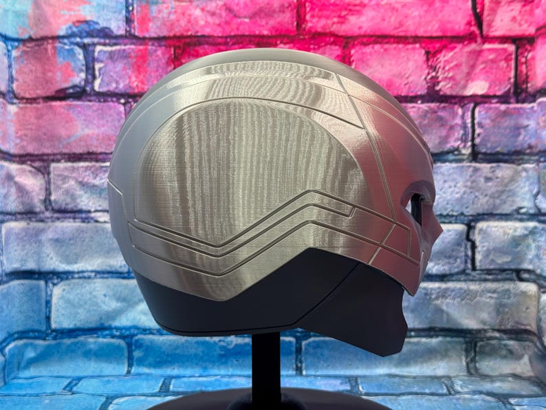 May include: A silver and black helmet with a brushed metal finish, displayed on a black stand. The helmet has a sleek, futuristic design with angular lines and a visor area. The background is a colorful brick wall.