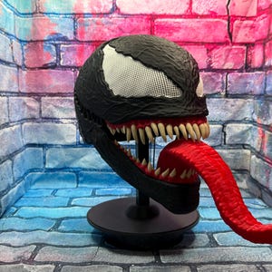 May include: A black Venom mask with white mesh eyes, sharp teeth, and a long red tongue. The mask is displayed on a black stand against a brick wall backdrop. The mask is a replica of the Marvel Comics character.
