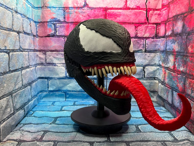 May include: A black Venom mask with white mesh eyes, sharp teeth, and a long red tongue. The mask is displayed on a black stand against a brick wall backdrop. The mask is a replica of the Marvel Comics character.
