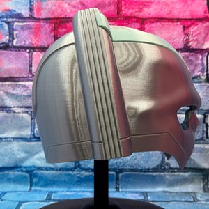 May include: A silver-colored helmet with a distinctive design, featuring a prominent vertical ridge on top and a visor-like opening. The helmet is displayed on a black stand against a colorful brick wall background.