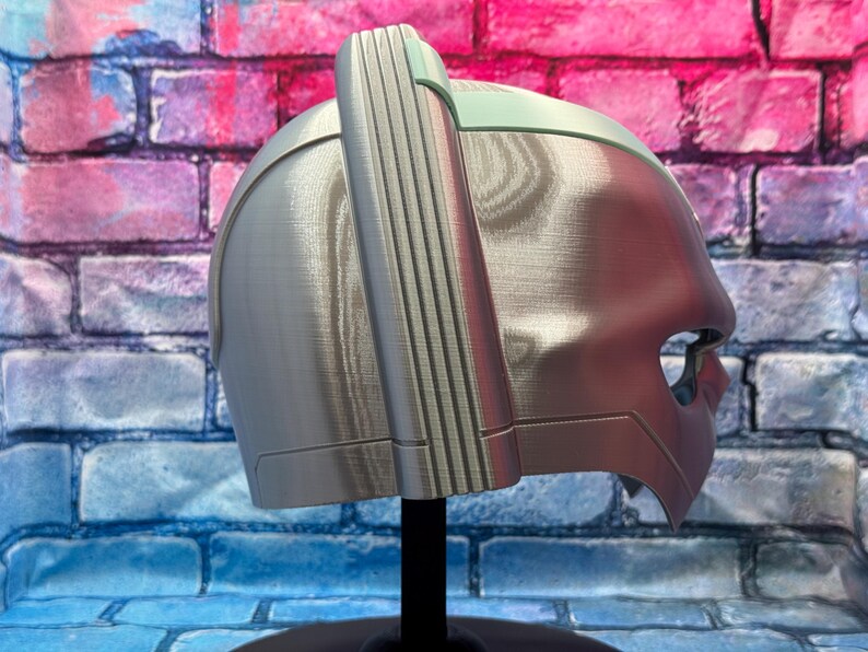 May include: A silver-colored helmet with a distinctive design, featuring a prominent vertical ridge on top and a visor-like opening. The helmet is displayed on a black stand against a colorful brick wall background.