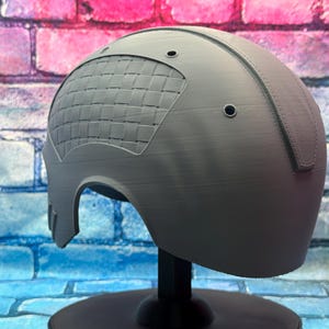May include: A gray helmet with a textured surface and a woven pattern on the side. The helmet has two small circular holes and is displayed on a black stand against a brick wall background.