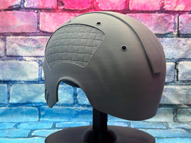 May include: A gray helmet with a textured surface and a woven pattern on the side. The helmet has two small circular holes and is displayed on a black stand against a brick wall background.