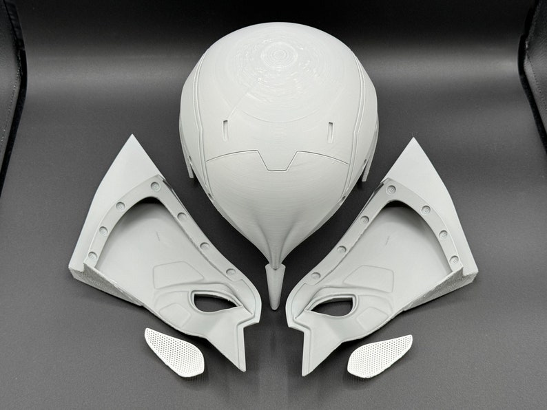 Puede incluir: Un conjunto de cinco piezas de casco de pl&aacute;stico gris, que incluye una pieza principal de casco con un dise&ntilde;o circular, dos piezas laterales y dos piezas m&aacute;s peque&ntilde;as. Las piezas est&aacute;n dispuestas sobre una superficie gris.