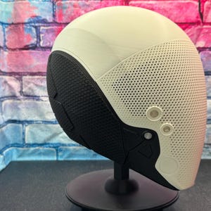 May include: A futuristic-looking helmet with a cream-colored top and a perforated, off-white front section. The lower portion is black with a textured pattern. Two circular, light-colored accents are visible on the front. The helmet is mounted on a black stand.