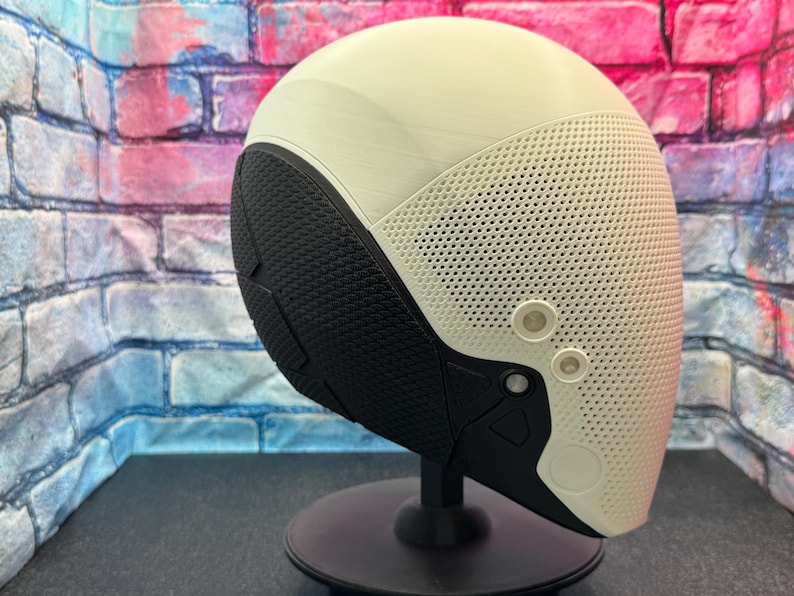 May include: A futuristic-looking helmet with a cream-colored top and a perforated, off-white front section. The lower portion is black with a textured pattern. Two circular, light-colored accents are visible on the front. The helmet is mounted on a black stand.