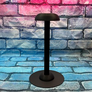 May include: A black hat stand with a round base and a tall, slender pole. The top of the stand has a curved, hat-shaped rest. The stand is made of metal and is designed to hold a hat. The background is a brick wall.