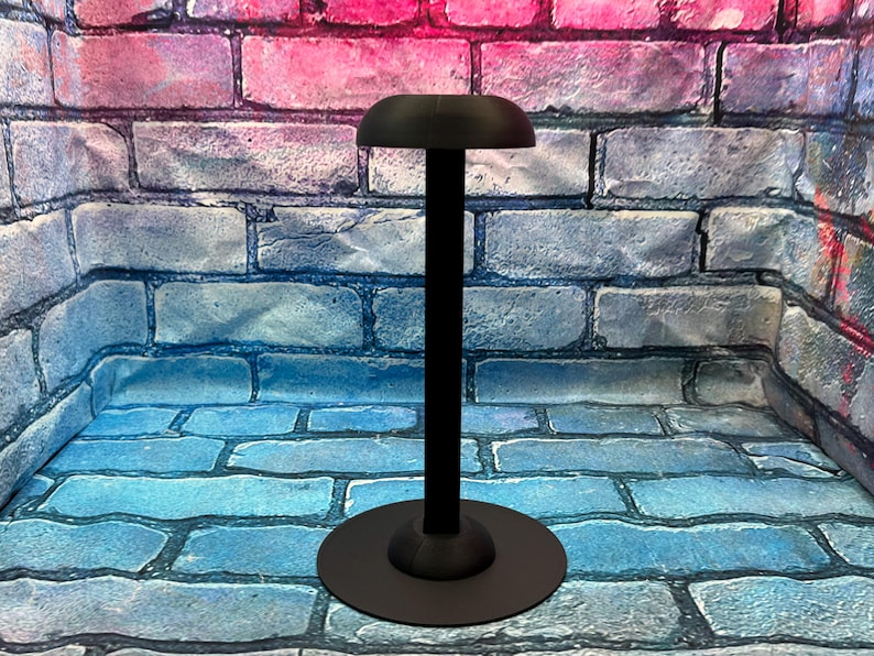 May include: A black hat stand with a round base and a tall, slender pole. The top of the stand has a curved, hat-shaped rest. The stand is made of metal and is designed to hold a hat. The background is a brick wall.