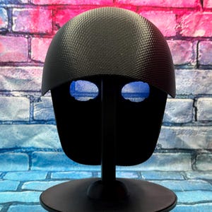 May include: A black helmet with a textured, carbon fiber-like pattern on the dome and a smooth, solid black faceplate featuring two distinct, horizontal, blue-tinted mesh eye openings, mounted on a black circular display stand.
