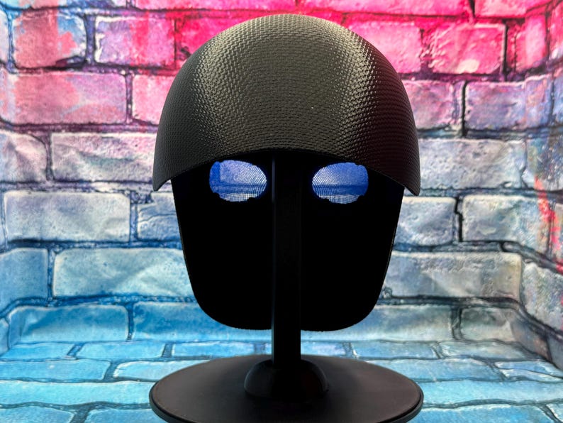 May include: A black helmet with a textured, carbon fiber-like pattern on the dome and a smooth, solid black faceplate featuring two distinct, horizontal, blue-tinted mesh eye openings, mounted on a black circular display stand.
