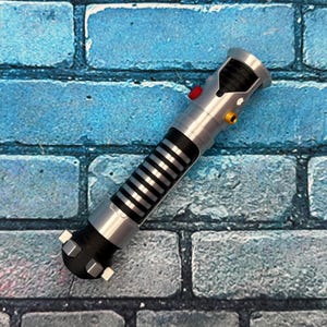 May include: A silver and black lightsaber prop with a red and yellow button detail. The prop has a black grip with silver stripes and is set against a blue brick wall.
