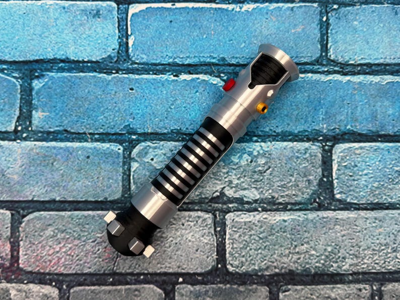 May include: A silver and black lightsaber prop with a red and yellow button detail. The prop has a black grip with silver stripes and is set against a blue brick wall.