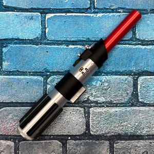 May include: A replica lightsaber with a red blade and a silver and black hilt. The hilt features detailed buttons and a ribbed grip. The lightsaber is set against a blue brick wall.