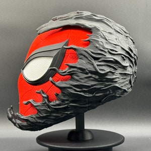 Puede incluir: Una m&aacute;scara de Spider-Man impresa en 3D en rojo y negro con un simbionte negro que cubre la parte superior y los lados de la m&aacute;scara. La m&aacute;scara est&aacute; sobre un soporte negro.