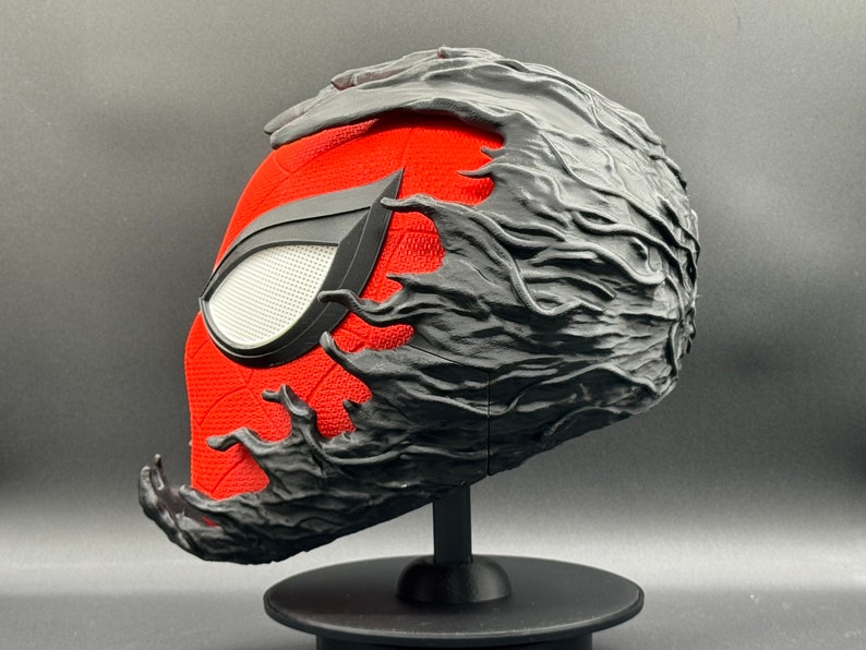 Puede incluir: Una m&aacute;scara de Spider-Man impresa en 3D en rojo y negro con un simbionte negro que cubre la parte superior y los lados de la m&aacute;scara. La m&aacute;scara est&aacute; sobre un soporte negro.