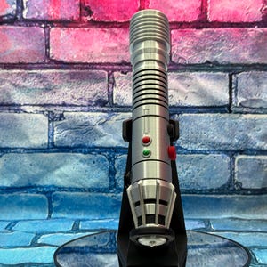 May include: A silver lightsaber replica with a black stand. The lightsaber has a ribbed handle, red and green buttons, and a detailed emitter. The background is a brick wall with pink, blue, and purple hues.