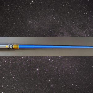 May include: A blue and silver lightsaber prop with gold accents. The hilt is silver, transitioning to a blue blade. The lightsaber is set against a starry background, suggesting a space or sci-fi theme.