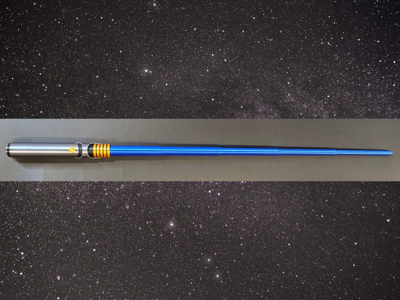 May include: A blue and silver lightsaber prop with gold accents. The hilt is silver, transitioning to a blue blade. The lightsaber is set against a starry background, suggesting a space or sci-fi theme.