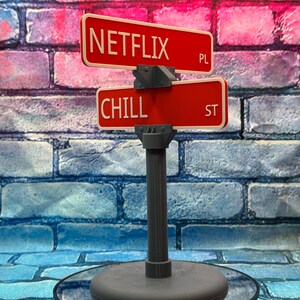 May include: A decorative street sign featuring two red signs with white lettering. The top sign reads "NETFLIX PL" and the bottom sign reads "CHILL ST". The sign is mounted on a gray post and base.