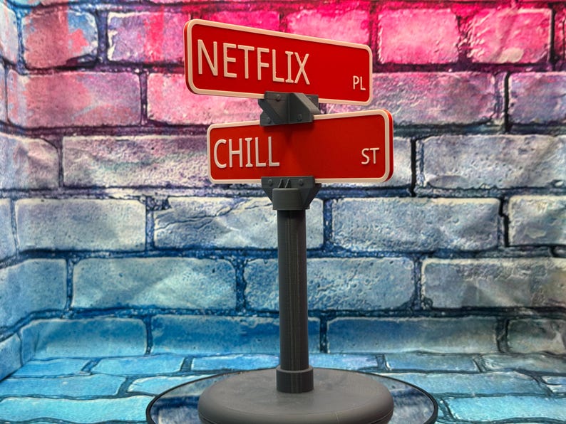May include: A decorative street sign featuring two red signs with white lettering. The top sign reads "NETFLIX PL" and the bottom sign reads "CHILL ST". The sign is mounted on a gray post and base.