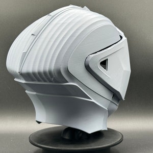 Puede incluir: Un casco blanco impreso en 3D con un dise&ntilde;o futurista. El casco tiene una visera y una superficie texturizada.