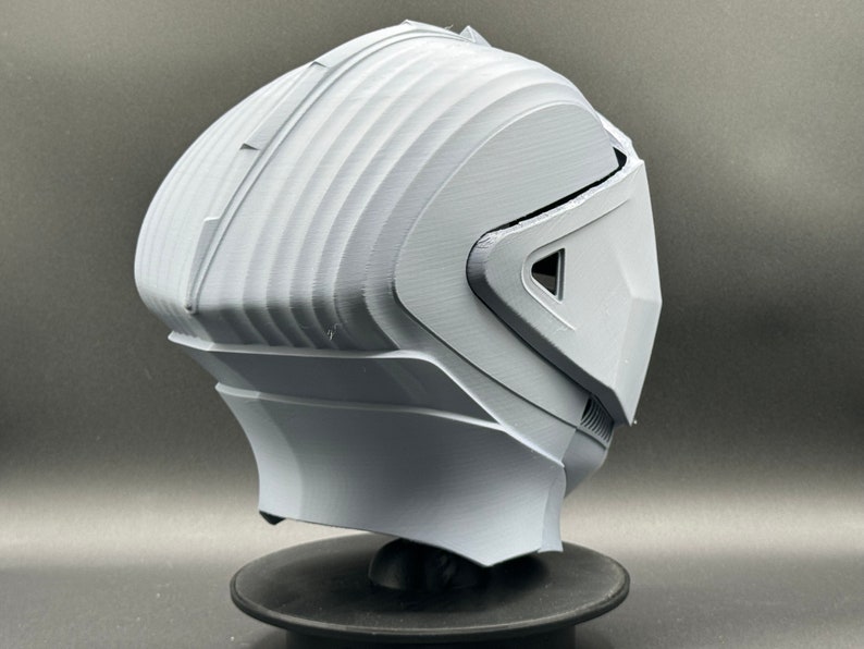 Puede incluir: Un casco blanco impreso en 3D con un dise&ntilde;o futurista. El casco tiene una visera y una superficie texturizada.