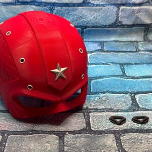 May include: A bright red helmet with a silver star emblem and silver rivets. The helmet has a diamond-patterned section on the side. Two black straps are next to the helmet. Superhero-themed cosplay.