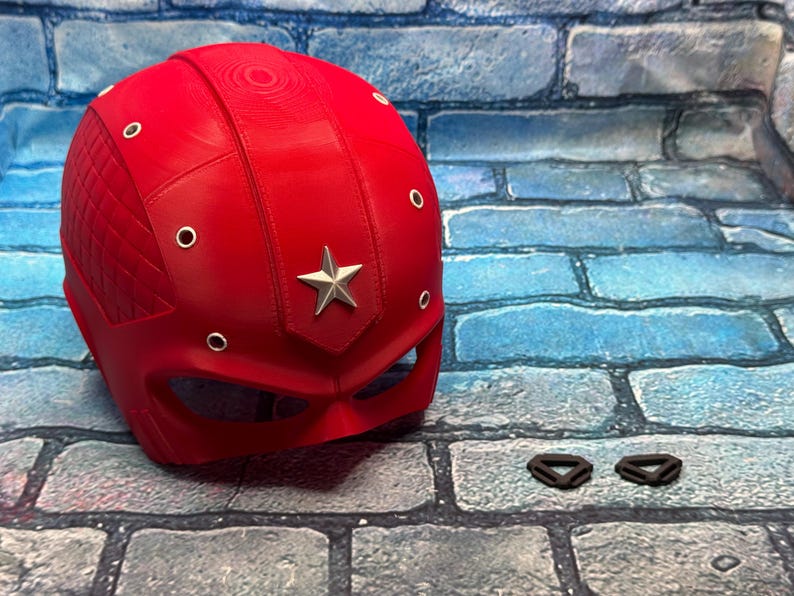 May include: A bright red helmet with a silver star emblem and silver rivets. The helmet has a diamond-patterned section on the side. Two black straps are next to the helmet. Superhero-themed cosplay.