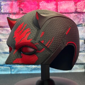 May include: A black and red helmet with a textured surface, featuring red accents and horns. The helmet has a mesh eye covering and is displayed on a black stand, showcasing a superhero or cosplay theme.
