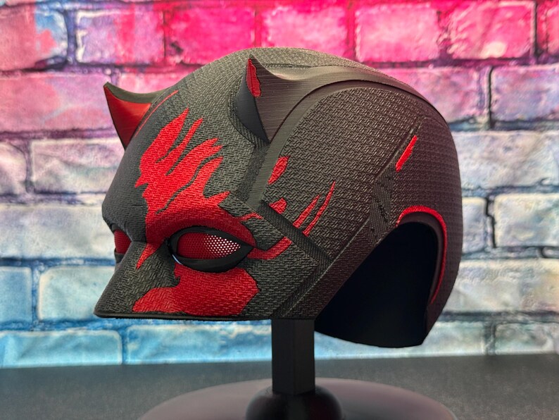 May include: A black and red helmet with a textured surface, featuring red accents and horns. The helmet has a mesh eye covering and is displayed on a black stand, showcasing a superhero or cosplay theme.