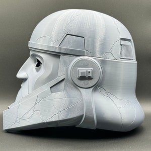 May include: A gray 3D printed helmet with a textured surface and a detailed design. The helmet has a visor and a side panel with a raised rectangular shape.