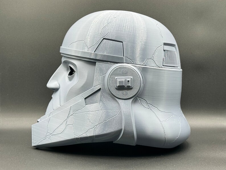 May include: A gray 3D printed helmet with a textured surface and a detailed design. The helmet has a visor and a side panel with a raised rectangular shape.