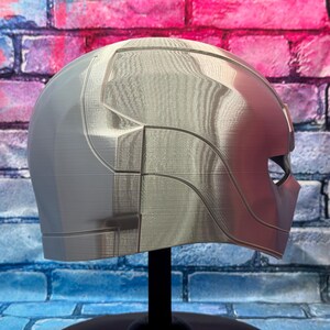 May include: A silver and red helmet with a geometric design. The helmet has a smooth, metallic finish and is displayed on a black stand. The design includes angular lines and a visor area. The background is a brick wall with blue, pink, and purple paint.