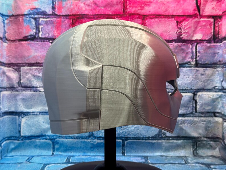 May include: A silver and red helmet with a geometric design. The helmet has a smooth, metallic finish and is displayed on a black stand. The design includes angular lines and a visor area. The background is a brick wall with blue, pink, and purple paint.