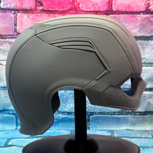 May include: A gray helmet with a futuristic design, featuring angular lines and textured details. The helmet is displayed on a black stand against a colorful brick wall background. The helmet has a visor area and a chin guard.