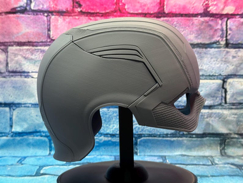 May include: A gray helmet with a futuristic design, featuring angular lines and textured details. The helmet is displayed on a black stand against a colorful brick wall background. The helmet has a visor area and a chin guard.