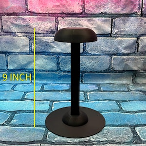 May include: A black helmet stand with a round base and a 9-inch (22.86 cm) tall central support. The top of the stand has a curved, helmet-shaped rest. The stand is designed to display a helmet.
