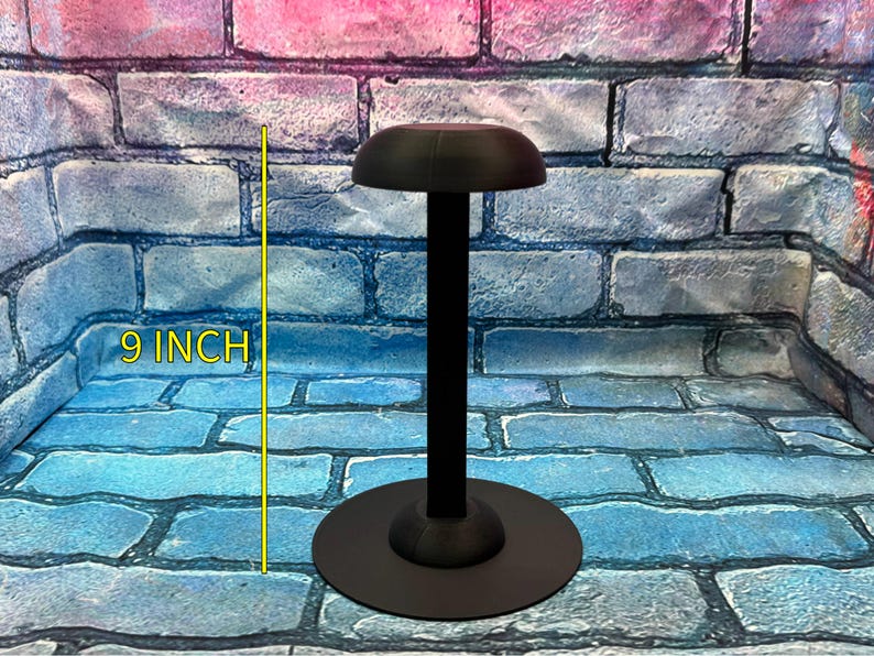 May include: A black helmet stand with a round base and a 9-inch (22.86 cm) tall central support. The top of the stand has a curved, helmet-shaped rest. The stand is designed to display a helmet.