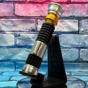 May include: A lightsaber prop in silver, gold, and black, presented on a black stand. The lightsaber has a ribbed black handle, silver details, and a gold section. The stand is angled, with a brick wall backdrop.