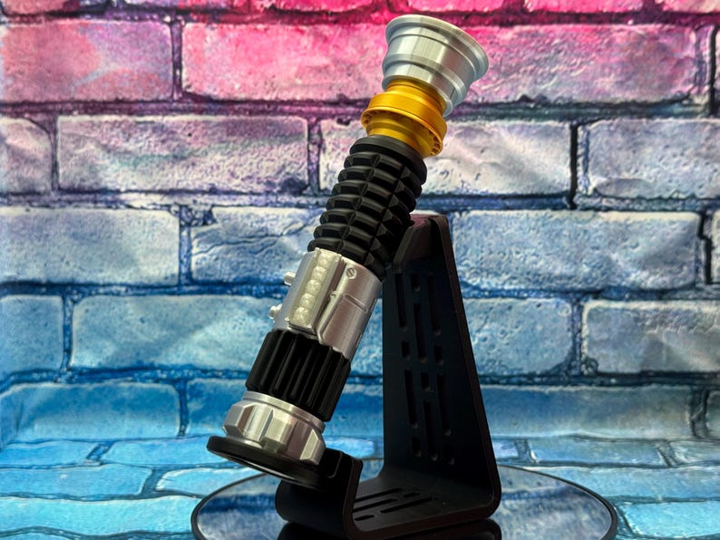 May include: A lightsaber prop in silver, gold, and black, presented on a black stand. The lightsaber has a ribbed black handle, silver details, and a gold section. The stand is angled, with a brick wall backdrop.