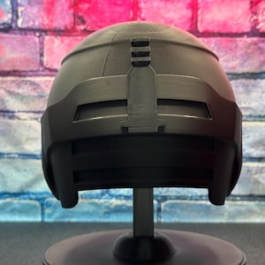 May include: A black helmet with a matte finish, mounted on a black stand. The helmet has a rounded top with a rectangular visor and a vertical bar detail. The background is a brick wall with pink, blue, and white paint.