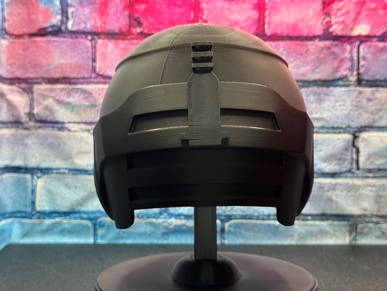 May include: A black helmet with a matte finish, mounted on a black stand. The helmet has a rounded top with a rectangular visor and a vertical bar detail. The background is a brick wall with pink, blue, and white paint.