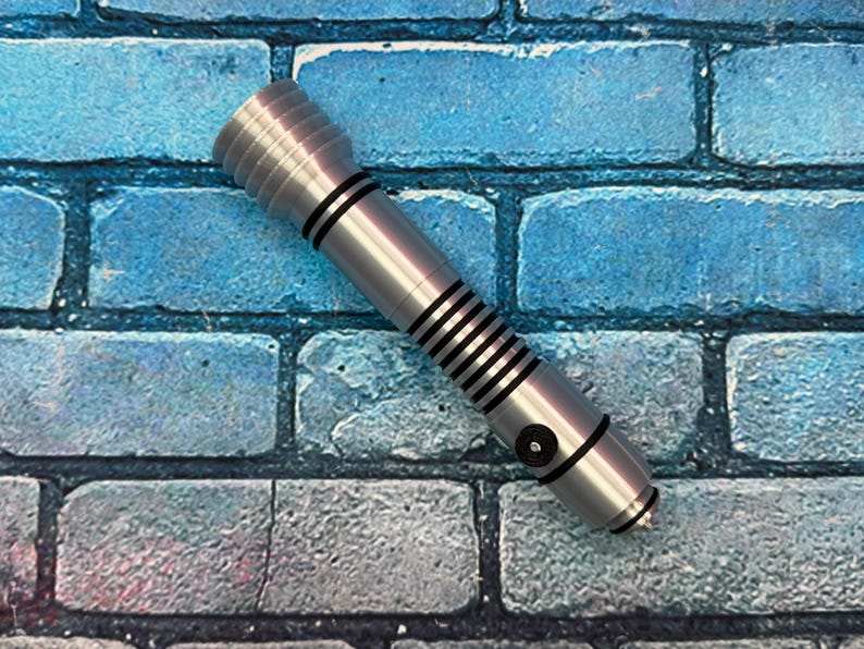 Puede incluir: Un accesorio de sable de luz plateado con detalles en negro, sobre un fondo de ladrillos azules. El sable de luz tiene un mango acanalado y rayas negras. El accesorio est&aacute; dise&ntilde;ado para parecerse a un arma futurista.